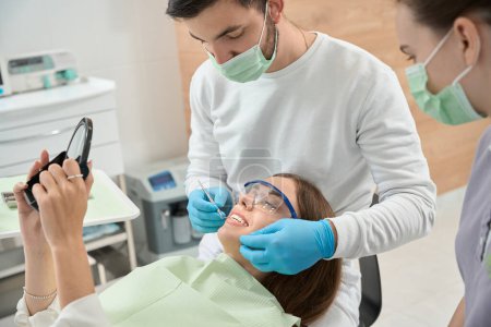 Foto de Mujer mirando en el espejo de la mano mientras el dentista revisa sus dientes anteriores con sonda dental en presencia de la enfermera - Imagen libre de derechos