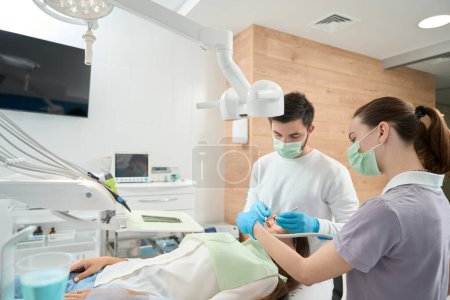 Téléchargez les photos : Médecin spécialisé dans les gants en nitrile inspectant la cavité buccale d'une patiente à l'aide d'instruments dentaires assistés par une infirmière - en image libre de droit
