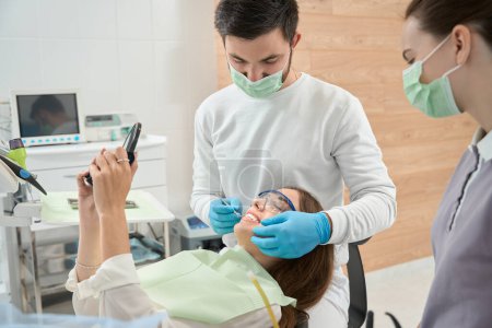 Téléchargez les photos : Patiente se regardant dans le miroir de la main tandis que la stomatologue vérifie ses dents supérieures avant en présence de l'infirmière - en image libre de droit