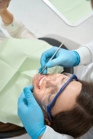 Photo for Female patient wearing bib and protective eyewear while dentist inspecting her front teeth - Royalty Free Image
