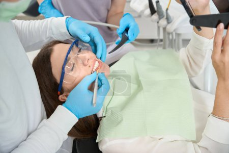 Téléchargez les photos : Patiente regardant dans le miroir de la main tandis que le dentiste enlever tartre entre ses dents assisté par l'infirmière - en image libre de droit
