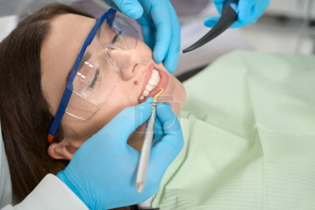 Foto de Cliente femenina en gafas protectoras acostada supina mientras dentista profesional quita el sarro entre sus dientes - Imagen libre de derechos