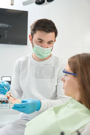 Foto de Dentista en mascarilla que muestra modelo de caries dental a cliente femenino sentado en silla - Imagen libre de derechos
