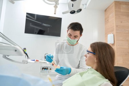 Téléchargez les photos : Stomatologue assis à côté du patient et pointant vers les caries sur le modèle de dent avec sonde dentaire - en image libre de droit