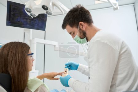 Photo for Stomatologist in face mask demonstrating dental caries model to serious female patient seated in chair - Royalty Free Image