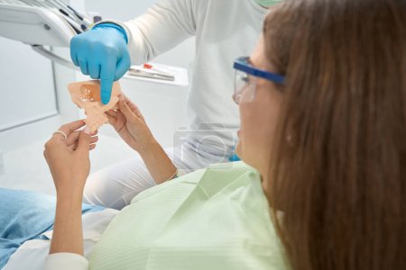Téléchargez les photos : Stomatologue en gants de nitrile pointant vers la pommette sur le modèle maxillaire dans les mains des patientes - en image libre de droit