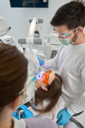 Téléchargez les photos : Hygiéniste dentaire expérimenté visant la lampe de blanchiment des dents de lumière LED à l'émail du client en présence de l'assistant - en image libre de droit