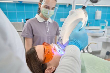 Photo for Doctor hand in nitrile glove directing LED light teeth whitening lamp at young woman tooth enamel - Royalty Free Image