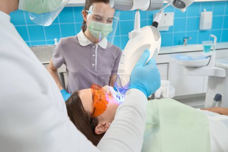 Téléchargez les photos : Hygiéniste dentaire professionnel utilisant la lampe de blanchiment de dents de lumière de LED sur l'émail dentaire de la jeune femme - en image libre de droit
