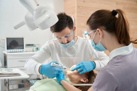 Téléchargez les photos : Stomatologue ciblée administrant une anesthésie locale à une femme utilisant une seringue dentaire aidée par une infirmière - en image libre de droit