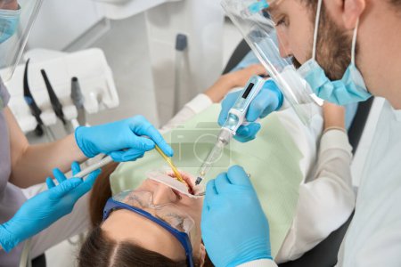 Téléchargez les photos : Dentiste expérimenté engourdissant la bouche du patient avant le traitement des dents assisté par une infirmière - en image libre de droit
