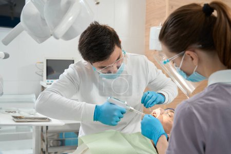 Photo for Experienced male dentist injecting local anesthetic to patient assisted by female nurse - Royalty Free Image