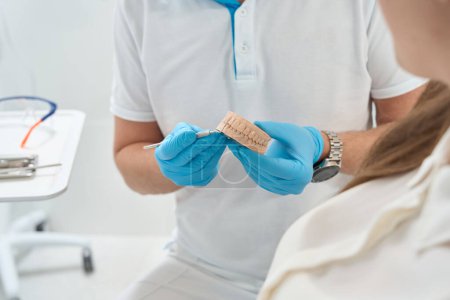 Foto de Foto recortada del médico en guantes de nitrilo apuntando con instrumento al modelo de dientes al paciente - Imagen libre de derechos