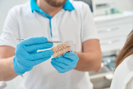 Foto de Foto recortada del dentista en guantes de nitrilo apuntando con herramienta médica al modelo de dientes al paciente - Imagen libre de derechos