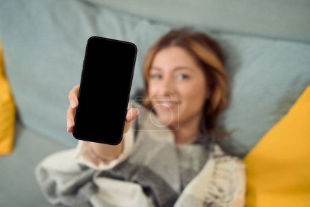 Foto de Smiling joyous female lying in bed and holding smartphone in front of camera - Imagen libre de derechos