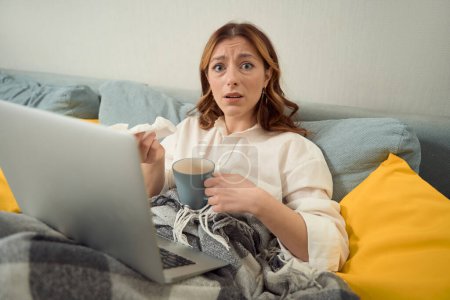 Photo for Upset young woman seated at laptop in bed holding handkerchief and mug in hands - Royalty Free Image