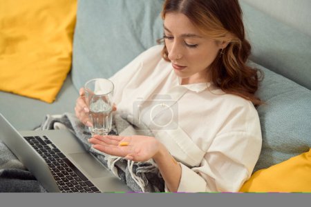 Téléchargez les photos : Sick lady sitting in bed with glass filled with water staring at drug capsule in her hand - en image libre de droit