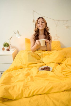 Photo for Smiling happy woman with mug of coffee in hands sitting in bed - Royalty Free Image
