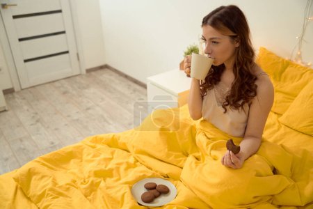 Foto de Female seated in bed with oatmeal cookie in hand drinking coffee in ceramic mug - Imagen libre de derechos