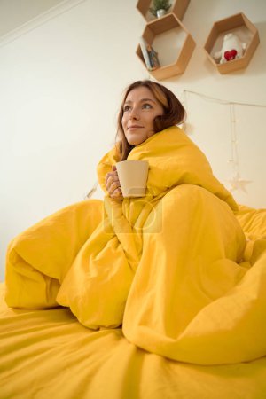 Téléchargez les photos : Dreamy lady wrapped in blanket holding mug of coffee and looking at ceiling - en image libre de droit