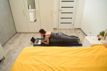 Foto de Fit woman performing forearm plank in front of laptop on yoga mat in bedroom - Imagen libre de derechos