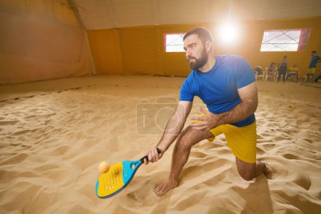 Foto de Serious tennis player in shorts and t-shirt knelt on one knee on the sand indoors while his racket touches the ball - Imagen libre de derechos
