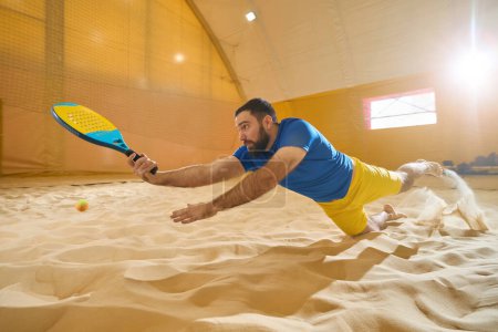 Téléchargez les photos : Concentrated tennis player firmly gripping the racket with his hand while falling on the sand during a sports game - en image libre de droit