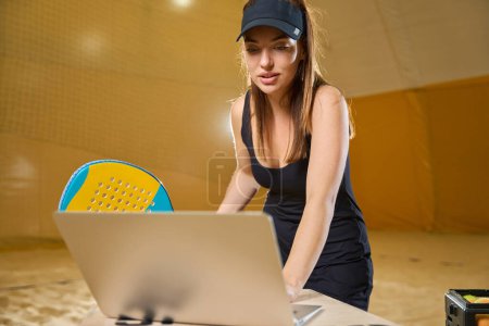 Foto de Focused female in sports uniform studies information on the laptop screen while holding a tennis racket in hands - Imagen libre de derechos