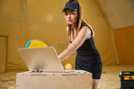 Foto de Focused female in sports uniform using gadget while holding tennis racket in playing hall - Imagen libre de derechos