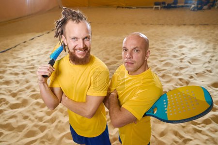 Foto de Confident men in yellow t-shirts stand on the sand indoors while holding wooden rackets in their hands - Imagen libre de derechos