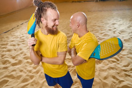 Téléchargez les photos : Cheerful beach tennis partners smiling at each other while standing side by side on sand indoors in shorts and t-shirt - en image libre de droit