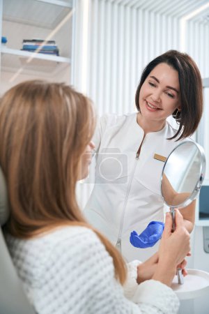 Foto de Young woman with mirror is located in a chair at the reception of beautician, specialist communicates kindly with the patient - Imagen libre de derechos