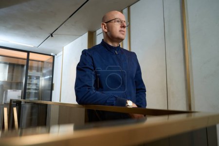 Téléchargez les photos : Bald middle-aged man stands leaning on the railing, there is a lot of light and glass around - en image libre de droit
