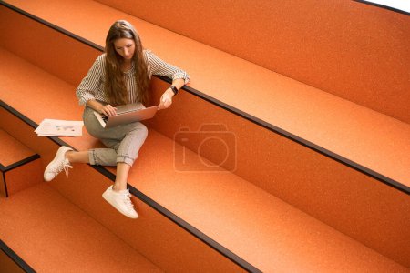 Foto de Top view of pretty young woman using laptop while sitting on steps, copy space - Imagen libre de derechos