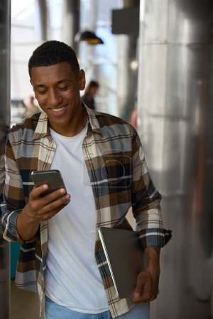 Téléchargez les photos : Happy African American guy reading the message on phone while holding laptop - en image libre de droit