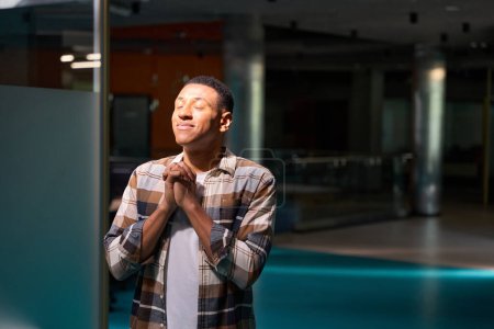 Foto de Smiling young African American man praying in the office while closing your eyes - Imagen libre de derechos