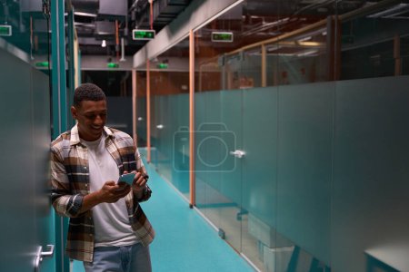Foto de Happy African American man typing on mobile phone while standing in the lobby in the office - Imagen libre de derechos