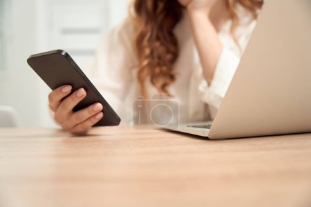 Foto de Cropped photo of woman sitting at laptop with mobile phone in hand - Imagen libre de derechos