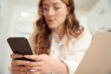 Photo for Portrait of serious focused woman sitting at laptop and texting on cellphone - Royalty Free Image
