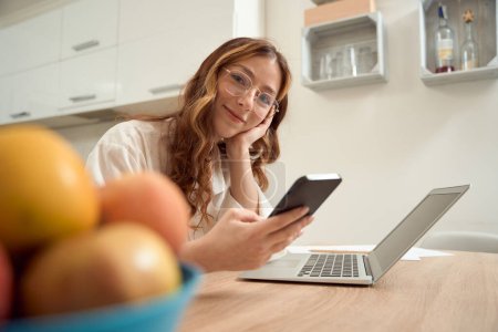 Photo for Smiling dreamy lady sitting at kitchen table with smartphone in hands - Royalty Free Image