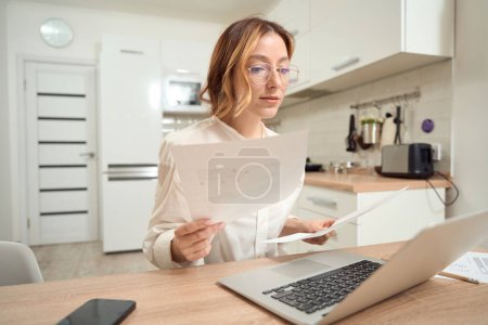 Photo for Concentrated lady sitting at kitchen table with bills in hands and staring at laptop screen - Royalty Free Image