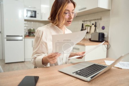 Photo for Focused young woman seated at kitchen table with documents in hands looking at laptop screen - Royalty Free Image