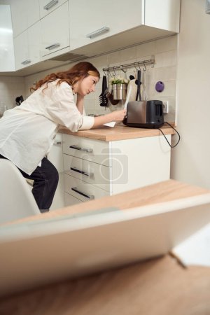 Photo for Puzzled female leaning on kitchen counter and staring at bill in her hand - Royalty Free Image