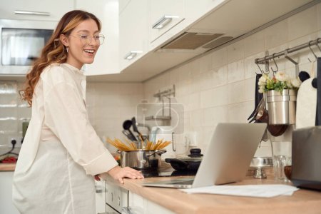 Foto de Smiling pleased young woman leaning on kitchen counter while looking at laptop screen - Imagen libre de derechos
