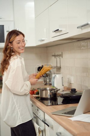Foto de Smiling woman standing at kitchen counter with spaghetti in hands and looking at laptop screen - Imagen libre de derechos