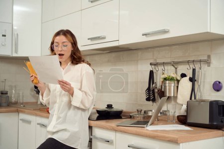 Téléchargez les photos : Focused woman with spaghetti in hand leaning against kitchen counter and looking at sheet of paper - en image libre de droit