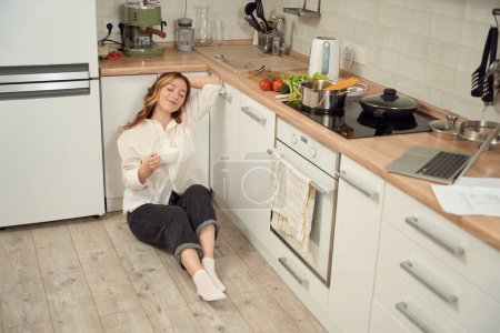 Foto de Smiling happy housewife seated with her eyes closed on kitchen floor holding ceramic cup in hands - Imagen libre de derechos