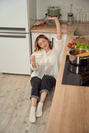 Foto de Pleased lady sitting with her eyes closed on wooden kitchen floor and holding coffee cup - Imagen libre de derechos