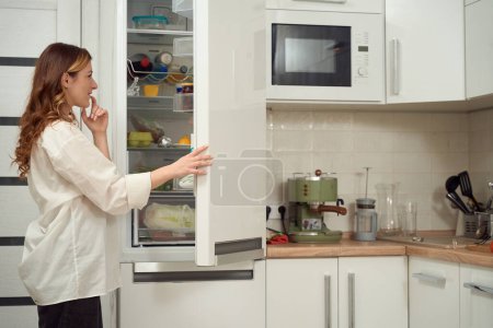 Foto de Side view of smiling woman standing in kitchen and looking on shelves with food in open refrigerator - Imagen libre de derechos