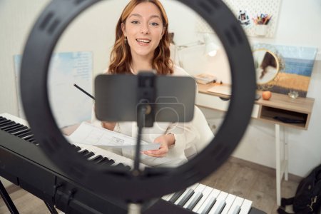 Foto de Cheerful synthesizer player holding sheet music and pencil in hands while looking into cellphone camera - Imagen libre de derechos